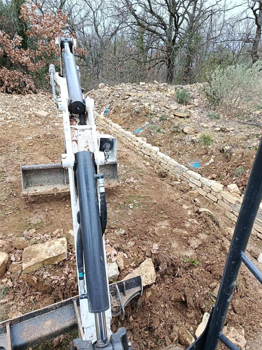 Bras de mini-pelle en cours de terrassement au pied d'un muret